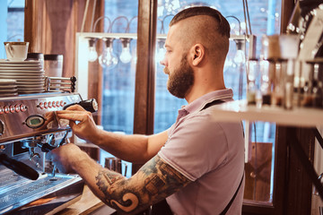 Handsome tattooed barista with stylish beard and hairstyle working on a coffee machine in a coffee shop or restaurant