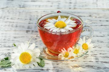 Chamomile tea. Cup of tea with chamomile, on which there is a ladybug, close-up, illuminated by sunlight, on a white wooden table.