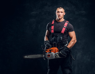 Handsome young lumberjack wearing protective clothes posing with a chainsaw in a dark studio