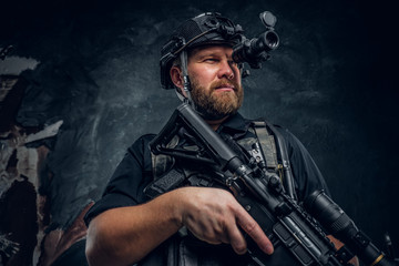 Bearded special forces soldier or private military contractor holding an assault rifle and observes the surroundings in night vision goggles. Studio photo against a dark textured wall