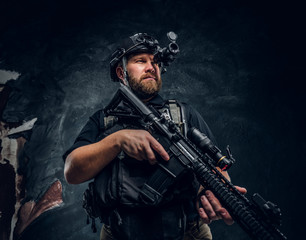 Fototapeta premium Bearded special forces soldier or private military contractor holding an assault rifle and observes the surroundings in night vision goggles. Studio photo against a dark textured wall
