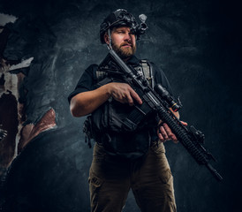 Bearded special forces soldier or private military contractor holding an assault rifle and observes the surroundings in night vision goggles. Studio photo against a dark textured wall