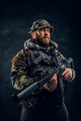 Brutal man in the military camouflaged uniform holding an assault rifle. Studio photo against a dark textured wall
