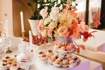 Candy bar with tasty desserts and appetizers at wedding reception. Decorated with flower compositions and candles. Sweet food, dessert, buffet, catering table, restaurant.