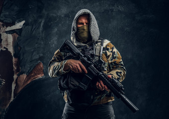 Special forces soldier in military uniform wearing mask and hood holding an assault rifle. Studio photo against a dark textured wall