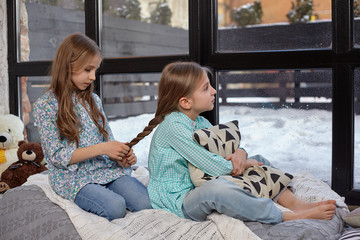 The image of two cute little sisters sitting on windowsill in peace and quiet
