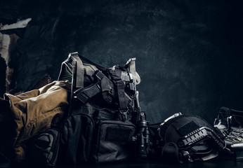 Military uniform and equipment. Body armor, gun, helmet, night vision goggles, boots. Studio photo against a dark textured wall