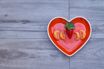 Front view of a heart-shaped dish with strawberries and tangerine inside, on a neutral gray floor