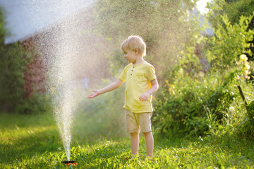 Funny little boy playing with garden sprinkler in sunny backyard