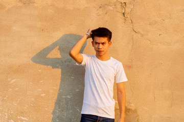 A young Thai boy is standing front of the dirty wall in the white shirt and shaping his hair.