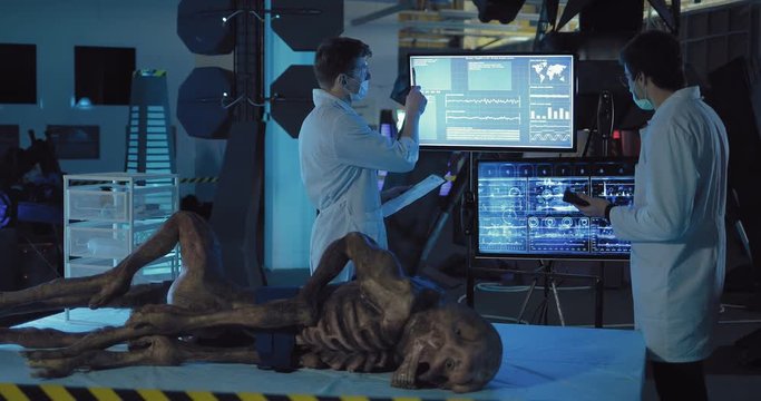 Scientists are experimenting with an alien from space. Men watch data on surveillance monitors. Alien lies on the table in the laboratory.