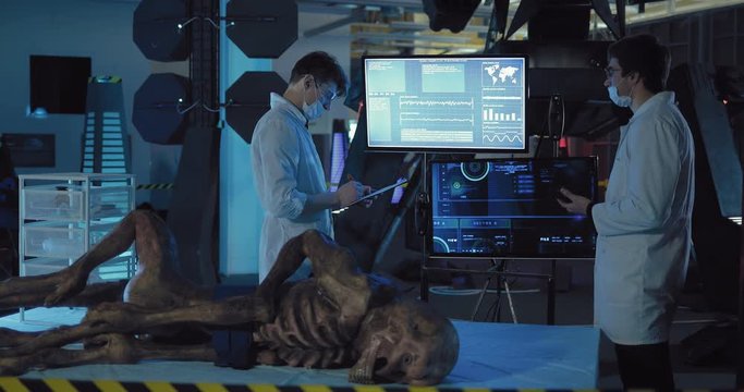 Young men scientists spend scientific experience in space laboratory, look at the data on the monitor. Alien lies in the technical unit.