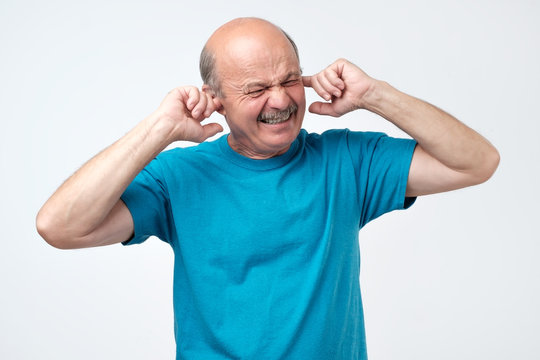 Senior hispanic bald guy plugging ears with fingers hearing loud sounds of music