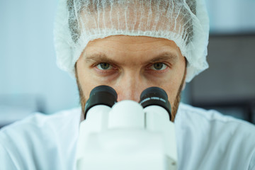 Close up of male face seriously looking at camera in medical cap. Young scientist with beard in lab coat looking under microscope. Crop of biologist working at modern laboratory on analysis.