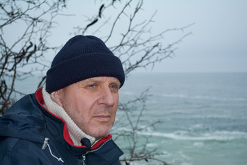 Realistic portrait of a middle-aged man with a thoughtful look in a sports jacket and hat against the background of the sea.