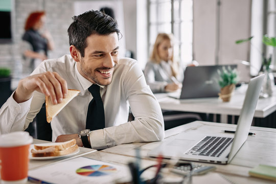 Happy businessman surfing the net on laptop while eating sandwich in the office. - Powered by Adobe