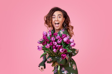 Cheerful and joyful young redhead woman in fashionable green dress being excited to get bouquet of spring yellow flowers tulips on women's day isolated over pink background. Gift Surprise Effect