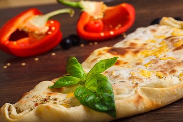 Close up calzone Italian pizza with melted cheese, tomatoes, fresh green herb basil leaves on a brown table decorated by mushrooms, black olives, red and green pepper and arugula leaves