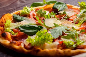 Close up pizza Italian with mozzarella, parmesan cheese, red tomatoes and fresh green basil, arugula and cabbage leaves on a brown table decorated by mozzarella, red pepper, cherry and battledore