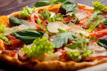 Close up pizza Italian with mozzarella, parmesan cheese, red tomatoes and fresh green basil, arugula and cabbage leaves on a brown table decorated by mozzarella, red pepper, cherry and battledore