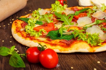 Close up pizza Italian with mozzarella, parmesan cheese, red tomatoes and fresh green basil, arugula and cabbage leaves on a brown table decorated by mozzarella, red pepper, cherry and battledore