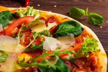 Close up pizza Italian with mozzarella, parmesan cheese, red tomatoes and fresh green basil, arugula and cabbage leaves on a brown table decorated by mozzarella, red pepper, cherry and battledore