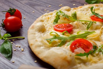 Vegetarian Italian pizza with melted mozzarella, parmezan cheese, red tomatoes and fresh green arugula leaves on a brown table decorated by mozzarella, red sweet pepper, cherry tomatoes and battledore