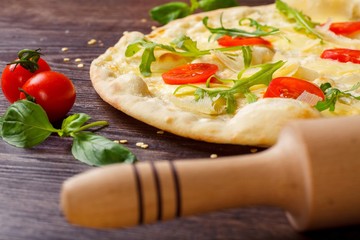 Vegetarian Italian pizza with melted mozzarella, parmezan cheese, red tomatoes and fresh green arugula leaves on a brown table decorated by mozzarella, red sweet pepper, cherry tomatoes and battledore