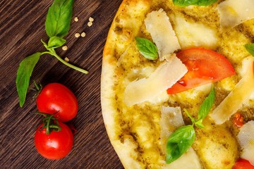 Close up vegetarian Italian pizza with melted mozzarella, parmesan cheese, red tomatoes and fresh green basil leaves on a brown table decorated by mozzarella, hot chili pepper, cherry and battledore