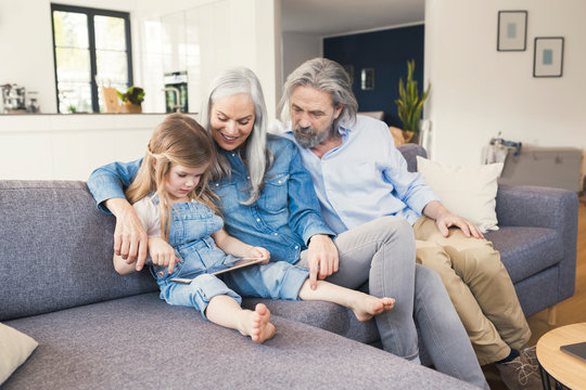 Grandparents and granddaughter sitting on couch, using tablet