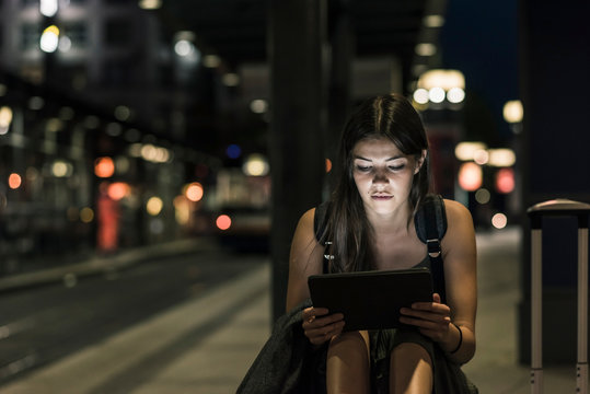 Portrait of young woman with headphones and tablet waiting at station by night