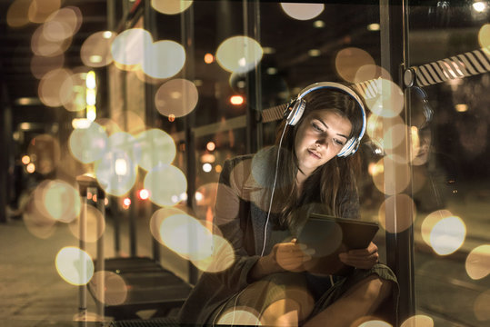 Portrait of young woman with headphones and tablet waiting at station by night