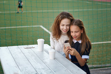 Two lovely teenage sisters have snack together next to school playground. Youngest girl holds cell...