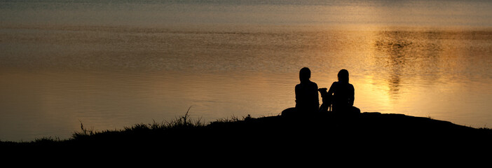 silhouette of two girls and a puppy sitting on the beach against a background of bright orange water space lit by the setting sun