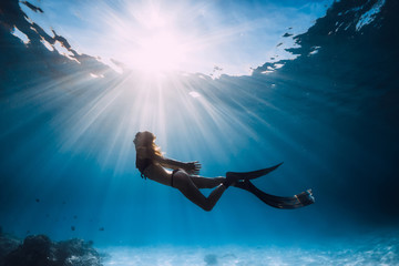Woman freediver with fins swim over sandy sea and sun rays underwater