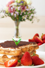  Tiramisu, homemade sweet cheesecake style dessert, on white plate embellished with fresh mint and a few real strawberries, cup of coffee, bowl with strawberries and flowers on white table. Closeup.
