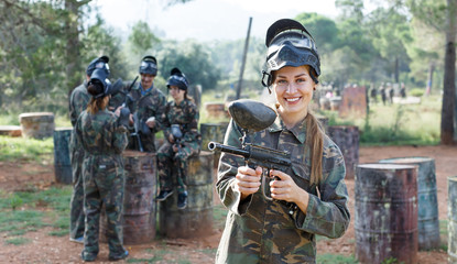 Fototapeta premium Young smiling woman in camouflage standing with gun before paintball match