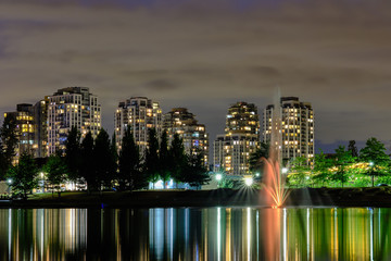 Night scape city view, Coquitlam, Greater Vancouver area, Canada. Fountain in lake and water reflections of modern buildings windows lights
