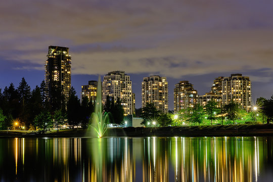 Night scape city view, Coquitlam, Greater Vancouver area, Canada. Fountain in lake and water reflections of modern buildings windows lights