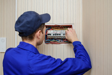 back view of a young eletrician in blue overall disassembling a electrical panel with fuses in a...
