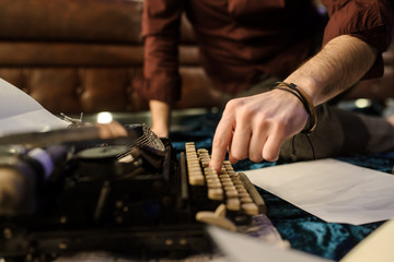 lateral view of man's hand typing on vintage typewriter on a piece of newspaper and dark blue velvet fabric on the floor in a dark room with light bulbs near a brown leather sofa