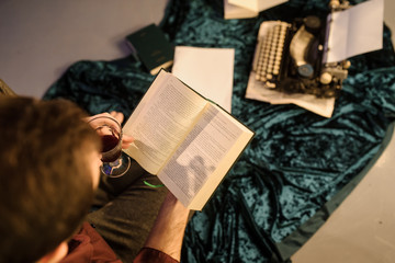 Fototapeta premium top view of man with a book in his hands sitting near a vintage typewriter on a dark blue velvet fabric on the floor with sheets of paper a glass of wine and books arround