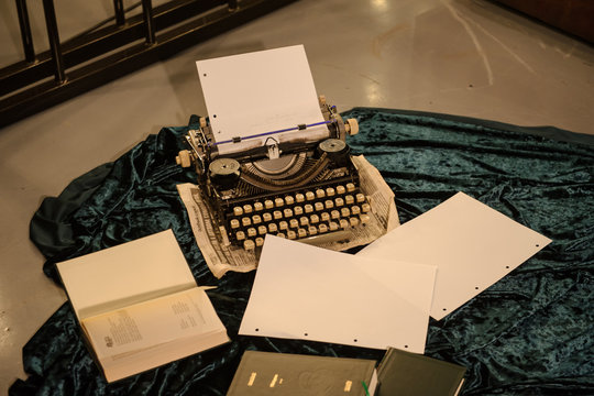 close up photo of a vintage typewriter on a piece of newspaper and dark blue velvet fabric on the floor with paper in it and other sheets of paper and a books arround