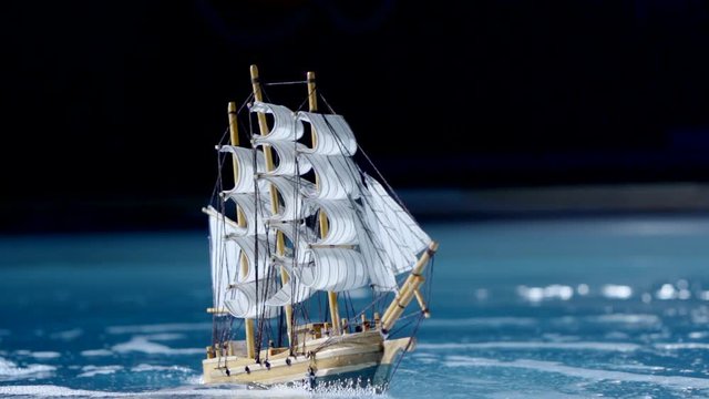 small toy ship with sails is floating on waves in pool, moving by underwater fountain