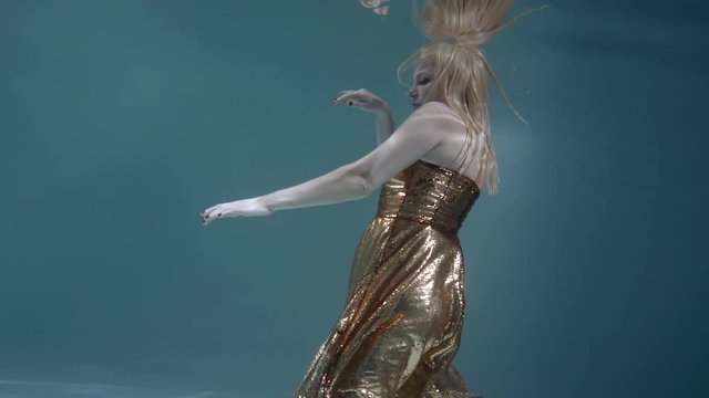 adult blonde woman is wearing golden dress is floating underwater in light blue background