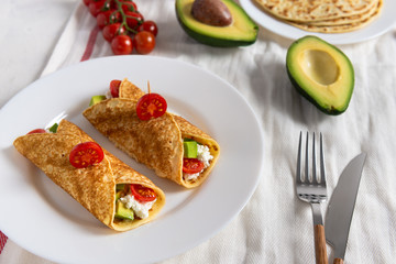Crepe pancakes with avocado, soft white cheese and cherry tomatoes on white plate. Cherry tomatoes, avocado and stack of crepes on side.