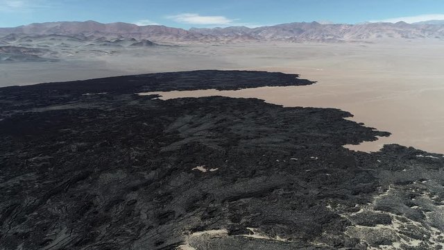 Flying over black bed of lava. Mountains, desertic landscape at background. Natural volcanic textures, patterns. From front view to senital. Antofagasta de la Sierra, Catamarca, Argentina