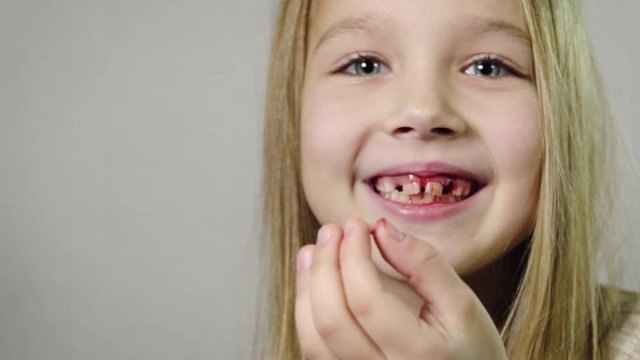 Close - up of a cute smiling girl, a child showing a torn milk Fang in her hand and a bleeding gums, jaw in a room with a white background. Tooth loss by age concept. 4K resolution