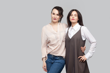 Portrait of two proud satisfied beautiful brunette best sisters in casual style standing, hugging each other and looking at camera with smile. indoor studio shot, isolated on grey background.