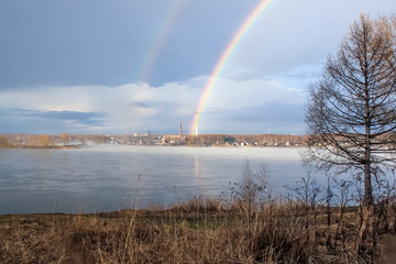 Peaceful spring misty landscape at sunrise after rain with rainbow and fog on the river Volga, in the background industrial plant with pipe. Сoncept of pollution and global warming.